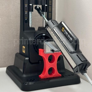 May include: A black and silver Work Sharp knife sharpener with a red support base. The sharpener has a silver sharpening rod and a black handle. The text "WORK SHARP" is visible on the handle.