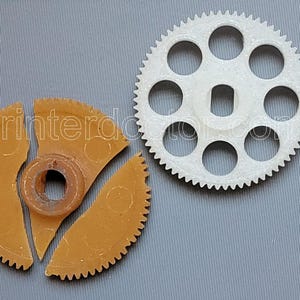May include: Two plastic gears, one white and one tan, are displayed against a gray background. The white gear is intact with circular holes. The tan gear is broken into three pieces. The image has the text "printerdoc.com" across the center.