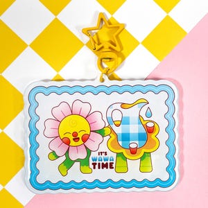 May include: A clear acrylic keychain with a cartoon illustration of a smiling flower and a pitcher of water. The keychain has a blue border and the text "IT'S WAWA TIME". It is attached to a yellow star-shaped clasp.