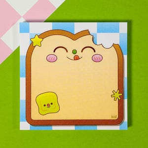 May include: A square notepad with a cartoon illustration of a slice of bread with a bite taken out. The bread has a smiling face, a star, and a butter square. The background is a blue and white checkered pattern.