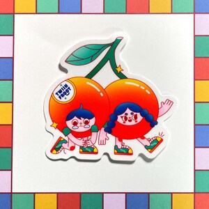 Puede incluir: Dos cerezas de dibujos animados con caras y piernas, que llevan zapatos verdes, se toman de la mano y caminan. Las cerezas son rojas y naranjas con tallos y hojas verdes. El texto "Twin Fruit" est&aacute; en una de las cerezas.
