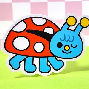 May include: A blue and red ladybug sticker with white spots and a winking face. The ladybug has two yellow antennas and is walking on four legs.