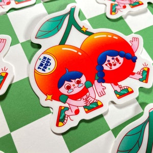 Puede incluir: Dos personajes de dibujos animados vestidos de cerezas con hojas verdes, tomados de la mano. Las cerezas son rojas y naranjas con contornos blancos. Los personajes tienen el pelo azul y sonr&iacute;en. El texto "Twin Fruit" est&aacute; escrito en la cereza de la izquierda.