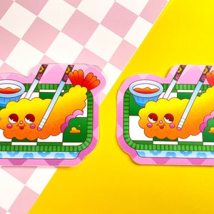 K&ouml;nnte beinhalten: Zwei rosa und gr&uuml;ne Cartoon-Aufkleber mit einer frittierten Garnele mit einem fr&ouml;hlichen Gesicht, Essst&auml;bchen und einer kleinen Sch&uuml;ssel mit orangefarbener Sauce. Die Aufkleber befinden sich auf einem rosa und wei&szlig;en karierten Hintergrund.