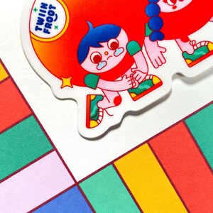 Puede incluir: Dos personajes de dibujos animados con ropa roja y verde y cabello azul. Los personajes se toman de la mano y sonr&iacute;en. Los personajes est&aacute;n sobre un fondo blanco con una pegatina que dice "Twin Froot".