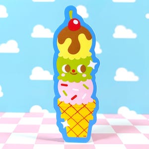 May include: A colorful ice cream cone sticker with three scoops: brown with a cherry, yellow, and pink with sprinkles. The cone is yellow with a grid pattern, and the sticker has a blue outline. The background is a blue sky with white clouds.