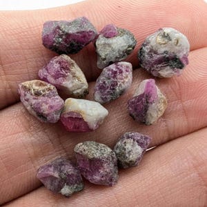 May include: A collection of raw ruby gemstones, displaying a mix of pink, red, and white hues. The rough stones have a natural, unpolished appearance, with visible crystalline structures. These gemstones are ideal for jewelry making or collecting.