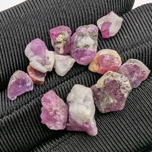 May include: A collection of raw ruby gemstones in various shapes and sizes. The stones display a range of colors, including deep pink, magenta, and white, with some showing hints of gold. The rough texture of the stones is visible.