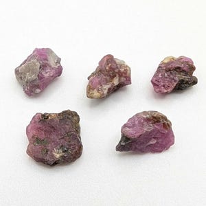 May include: Five rough-cut ruby gemstones in varying sizes and shapes. The raw stones display a deep pink and reddish-purple hue, with some areas showing a darker, almost black, coloration. The gemstones are set against a white background.