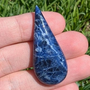 May include: A teardrop-shaped sodalite gemstone pendant. The polished stone features a deep blue color with white veining. The pendant is held in a person's hand, with green grass in the background.