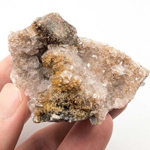 May include: A close-up of a mineral specimen featuring a cluster of translucent, cubic crystals. The crystals are a mix of clear and light brown hues, with areas of darker brown and gold. The specimen is held in a hand against a white background.