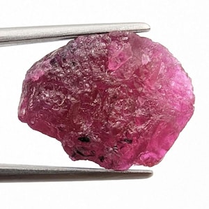 May include: A raw, unpolished gemstone with a deep magenta and pink hue. The stone features a jagged, crystalline surface with natural facets and irregular edges, held securely between the two metal prongs of a pair of silver-toned tweezers.