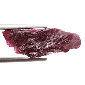 Puede incluir: Una piedra preciosa en bruto y sin pulir de tono magenta profundo y rojo oscuro. La piedra presenta una superficie irregular y dentada con facetas naturales y bordes translúcidos, sostenida horizontalmente por unas pinzas de metal plateado.