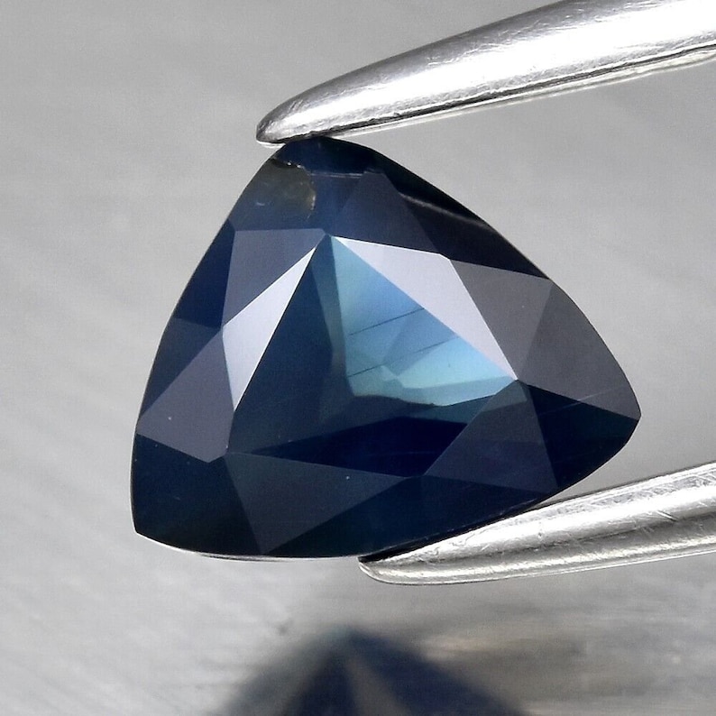 Puede incluir: Una piedra preciosa de zafiro triangular azul oscuro con un corte facetado. La piedra preciosa est&aacute; sujeta por unas pinzas.