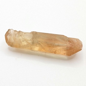 May include: A raw, natural crystal in shades of amber and light brown. The elongated, rectangular crystal has a rough, textured surface with visible internal patterns. The crystal is set against a white background.