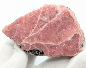May include: A raw rhodochrosite mineral specimen featuring a vibrant pink hue with intricate white crystalline veins running across the surface and a small patch of dark, textured material visible along the bottom edge of the stone.
