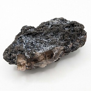 Peut inclure: Une formation rocheuse texturée, de couleur gris foncé, avec des cristaux translucides brun clair incrustés. La roche présente une surface rugueuse et poreuse avec des zones de scintillement blanc argenté, contrastant avec la couleur de base plus foncée.