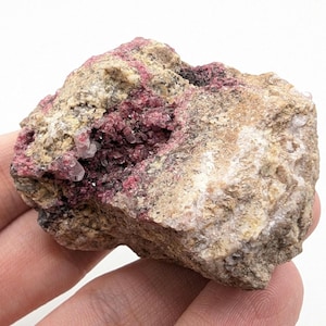 Puede incluir: Un espécimen mineral natural en bruto con una mezcla de colores. La roca tiene un exterior beige y bronceado con un interior rojo-rosa intenso. Pequeños cristales transparentes son visibles dentro de las grietas de la roca. La textura general es áspera y desigual.
