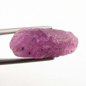 May include: A rough, uncut ruby gemstone, held by silver tweezers. The ruby is a vibrant pinkish-red colour with a textured surface and small black inclusions. The gemstone is displayed against a white background.