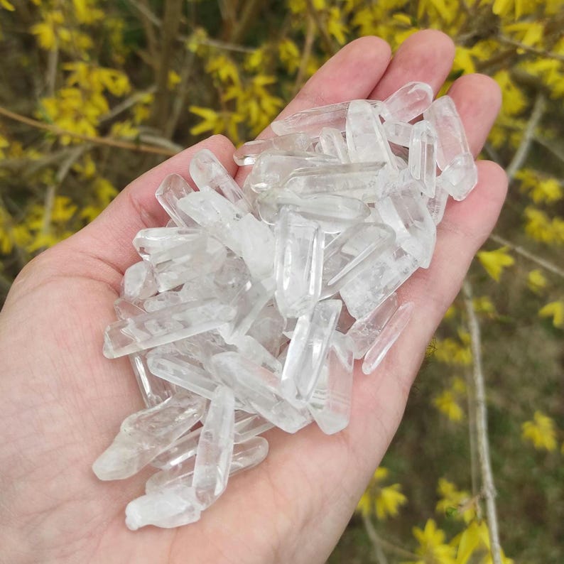 May include: A hand holding a large pile of clear quartz crystal points. The points are various sizes and shapes, and they are all very clear and shiny.
