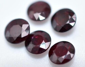 May include: Five round, faceted, dark red gemstones. The gemstones are arranged in a semi-circle on a white surface.