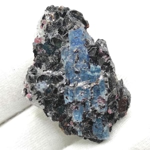 Peut inclure: Spécimen minéral brut avec un mélange de couleurs et de textures. La pierre présente une base sombre, presque noire, avec des touches de bleu, de blanc et de petits cristaux roses. La forme générale est irrégulière et rugueuse.