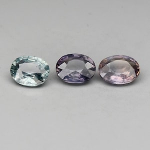 May include: Three oval-shaped gemstones. The left gemstone is a light blue-green color. The middle gemstone is a light purple color. The right gemstone is a light pink color.