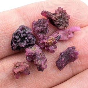 May include: A collection of rough, uncut ruby gemstones in varying shades of pink and red. The raw stones have a textured, crystalline appearance, with some showing darker, almost black, areas. The gemstones are held in a hand, showcasing their natural beauty.