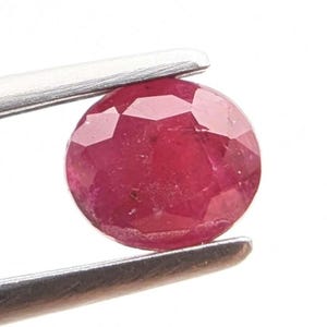 Peut inclure: Un rubis rond et facetté, maintenu par une pince argentée. La gemme rouge foncé présente des inclusions internes visibles. Le rubis est présenté sur fond blanc, mettant en valeur sa couleur et sa taille.