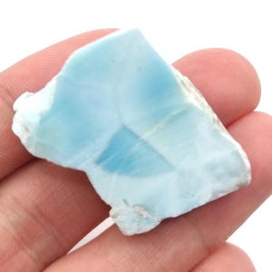 May include: A piece of larimar gemstone, a light blue stone with a marbled appearance. The stone has a mix of light and dark blue hues, with a rough, natural texture. The gemstone is held in a hand.