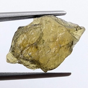 Puede incluir: Una gema en bruto, de color verde oliva, sujeta con pinzas. La piedra tiene una superficie texturizada y una forma irregular. El color es un verde amarillento apagado, con variaciones de tono. La gema parece ser un espécimen natural sin cortar.