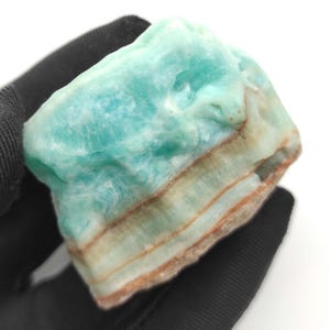 Pode incluir: Uma pedra natural bruta com uma aparência em camadas. A parte superior é de um turquesa vibrante, passando para tons mais claros e tons terrosos na base. A pedra tem uma superfície áspera e texturizada.
