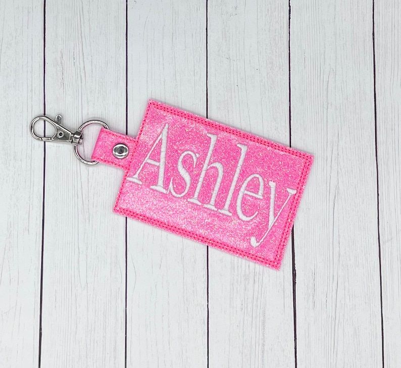 May include: Pink glitter keychain with a silver key ring and the name "Ashley" embroidered in white.