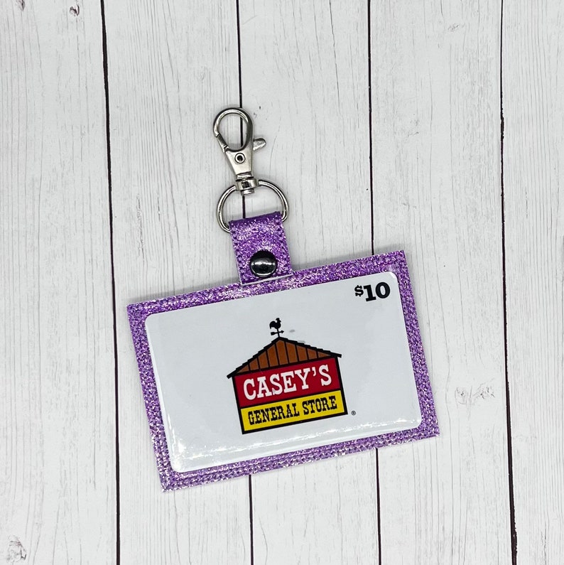 May include: Purple glitter gift card holder with a keychain. The gift card has a white background with a red and yellow logo that says "Casey's General Store" and the text "$10".