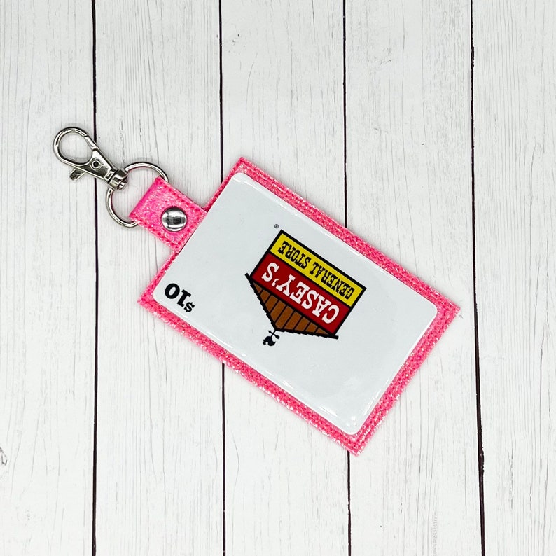 May include: Pink glitter keychain with a white Casey's General Store logo. The logo features a red roof and a yellow sign with the text "Casey's General Store". The keychain also has a silver metal ring and a $10 price tag.