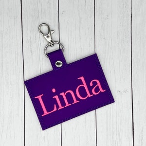 May include: A purple fabric key chain with a silver clip and the name "Linda" embroidered in pink.