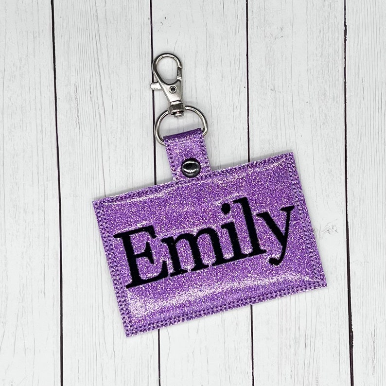 May include: Purple glitter keychain with a silver clip and the name "Emily" in black lettering.