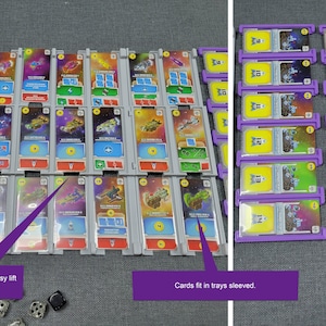 May include: A board game organiser with multiple trays and card holders. The trays are grey and purple, holding colourful cards with spaceship illustrations. Dice are scattered nearby. The organiser is designed for easy card access.