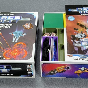 May include: A board game box set titled "Space Base Command Station" with a colourful outer box and an open inner box. The box art features spaceships and a fiery nebula. The set includes a field manual and game components.