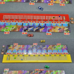 May include: A board game setup featuring four player boards in blue, red, yellow, and green. Each board has a numbered track and card slots. Dice and tokens are placed around the boards, and cards with spaceship designs are visible.