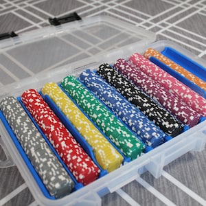 May include: A clear plastic case holding a set of poker chips. The chips are in various colors, including gray, red, yellow, green, blue, black, purple, pink, and orange. Each chip has a white speckled design. The case has a blue interior and a clear lid.