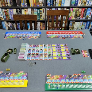May include: A board game laid out on a grey surface, with colourful game boards, cards, dice, and various game pieces. The game boards are arranged in rows with different coloured sections. A bookshelf filled with board games is in the background.