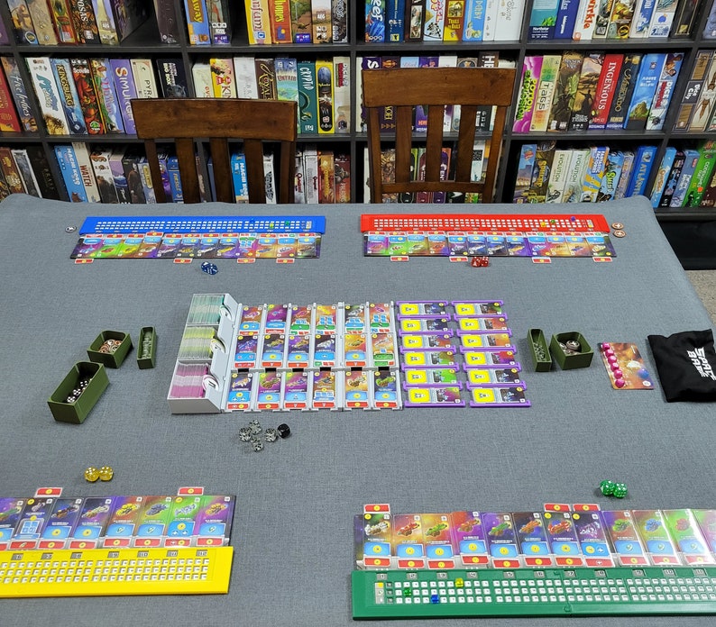 May include: A board game laid out on a grey surface, with colourful game boards, cards, dice, and various game pieces. The game boards are arranged in rows with different coloured sections. A bookshelf filled with board games is in the background.