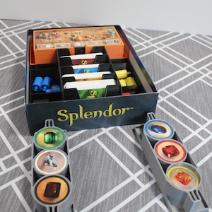 May include: A board game organizer for the game Splendor. The organizer is black and orange, with compartments for cards, tokens, and other game components. Two gray token holders are in the foreground, each holding three round tokens with gem illustrations. The game's name is printed on the box.