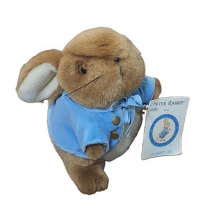 May include: A brown plush rabbit toy wearing a light blue jacket with two gold-colored buttons and a tie at the neck. The rabbit has long ears with white inner lining and black whiskers. A white tag attached reads Peter Rabbit from The World of Peter Rabbit.
