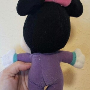 Pu&ograve; includere: Un peluche con testa nera, grandi orecchie rotonde e un fiocco rosa sulla parte superiore. Il giocattolo indossa una tutina viola con polsini color ottanio sulle maniche e mani di peluche bianche. Il tessuto ha una consistenza morbida e cuciture.