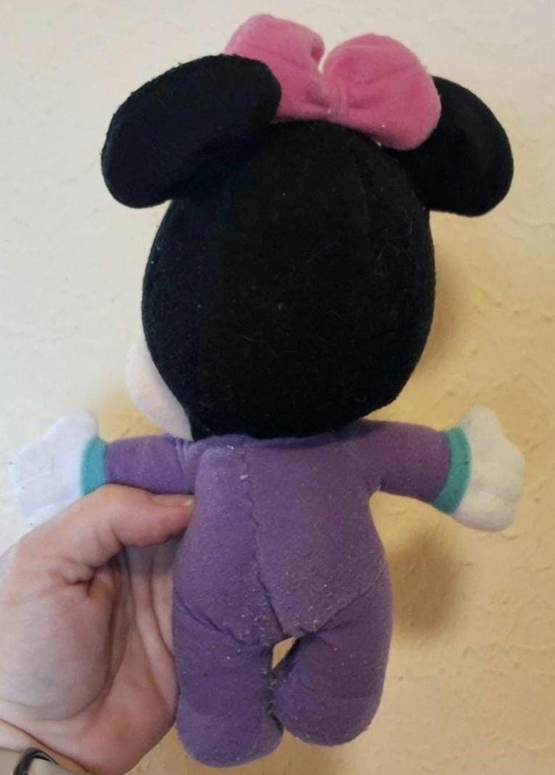 Pu&ograve; includere: Un peluche con testa nera, grandi orecchie rotonde e un fiocco rosa sulla parte superiore. Il giocattolo indossa una tutina viola con polsini color ottanio sulle maniche e mani di peluche bianche. Il tessuto ha una consistenza morbida e cuciture.