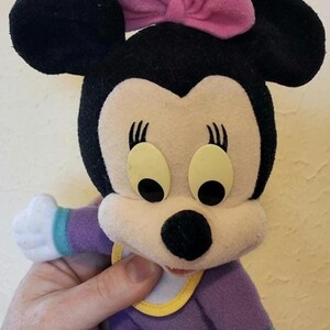 Pu&ograve; includere: Un peluche di un personaggio topo dei cartoni animati con orecchie nere, un fiocco in tessuto rosa e grandi occhi ovali. Il personaggio indossa un completo viola con maniche bianche, polsini color ottanio e una bavaglino bianco con bordo giallo.