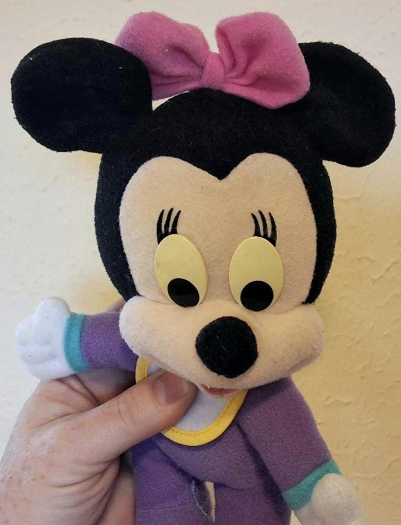 Pu&ograve; includere: Un peluche di un personaggio topo dei cartoni animati con orecchie nere, un fiocco in tessuto rosa e grandi occhi ovali. Il personaggio indossa un completo viola con maniche bianche, polsini color ottanio e una bavaglino bianco con bordo giallo.