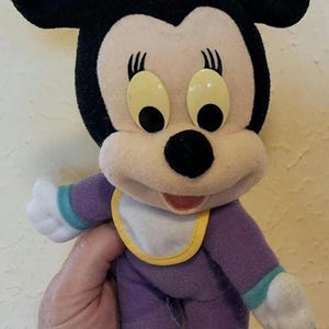 Pu&ograve; includere: Un peluche di un personaggio topo dei cartoni animati con orecchie nere, un fiocco rosa e grandi occhi gialli. Il personaggio indossa una tutina viola con polsini color ottanio e una bavaglino bianco con bordo giallo e mani bianche.